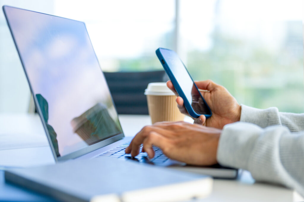 Close up of a Businessman working on a laptop computer and holding and looking at a mobile phone in the office. There are notepads on the desk. He is casually dressed